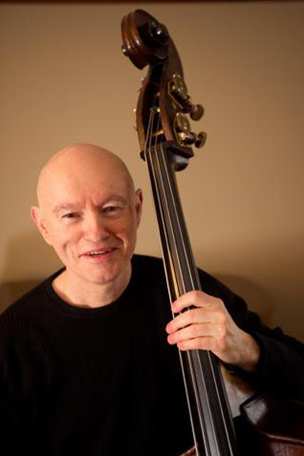 A person is shown playing an upright double bass in an indoor setting. The musician is wearing a dark sweater and is focused on the instrument, which is prominently featured in the foreground. The background is neutral, drawing attention to the double bass and the musician's hands. The lighting is warm, creating a cozy and intimate atmosphere.