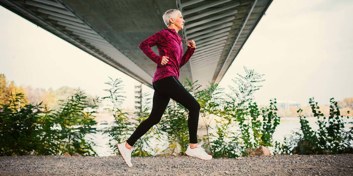 An older woman with short gray hair is jogging on a gravel path beneath a large bridge. She is dressed in athletic wear, including a red jacket and black leggings, and appears focused and energetic. The background features greenery and a river, suggesting an urban park or riverside setting.