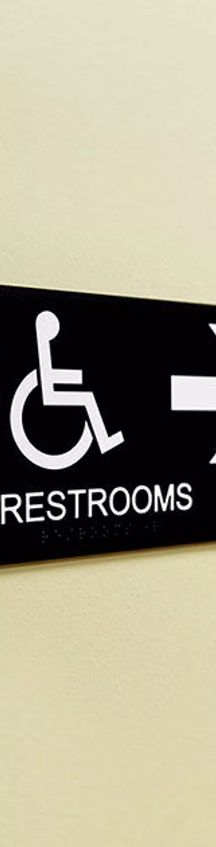 A black and white restroom sign is mounted on a light-colored wall, indicating the direction to accessible restrooms. The sign features icons for male, female, and wheelchair access, with a large arrow pointing right. The background shows a hallway with red walls and several closed doors, suggesting a public or commercial building. The word 'RESTROOMS' is clearly visible on the sign.