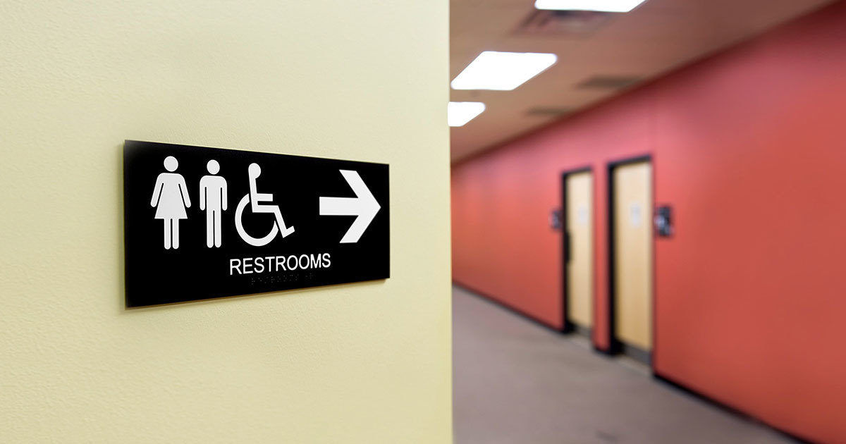 A black and white restroom sign is mounted on a light-colored wall, indicating the direction to accessible restrooms. The sign features icons for male, female, and wheelchair access, with a large arrow pointing right. The background shows a hallway with red walls and several closed doors, suggesting a public or commercial building. The word 'RESTROOMS' is clearly visible on the sign.