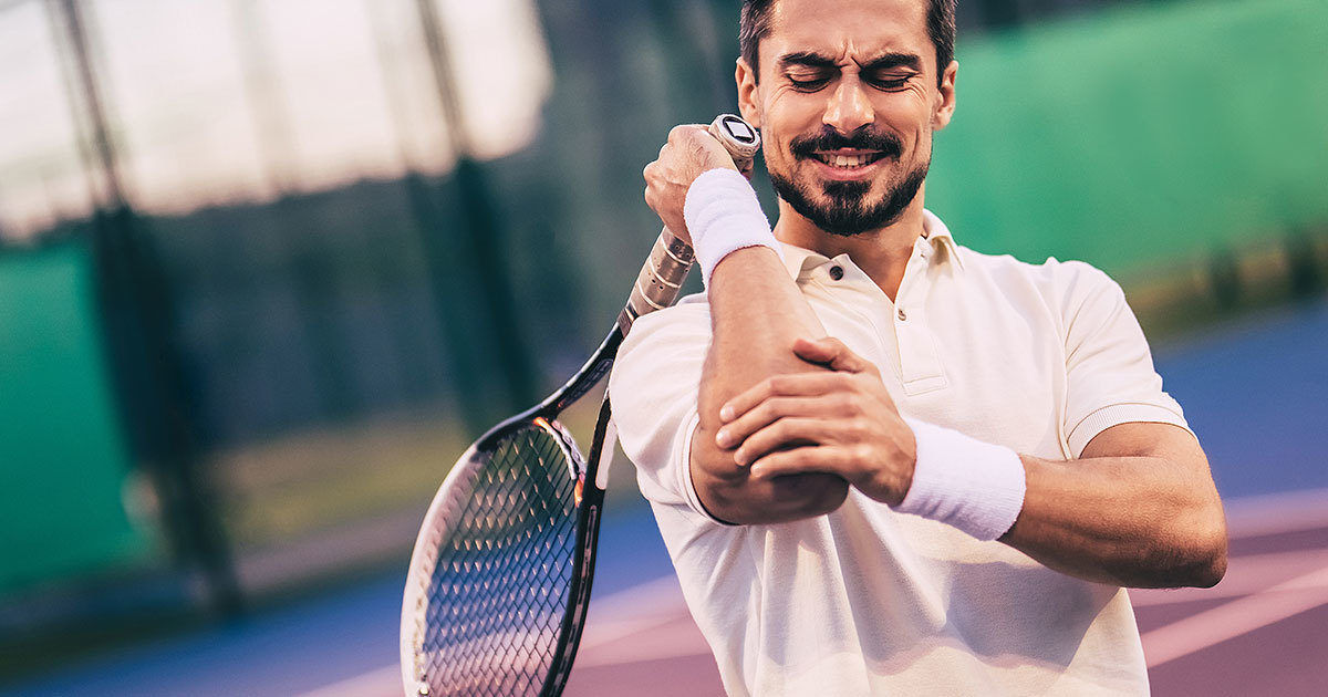 A male tennis player in a white polo shirt stretches his arm while holding a tennis racket. The scene is set on an outdoor tennis court with a blurred green and purple background. The player wears white wristbands and appears to be preparing for a match or practice. The image conveys a sense of focus and athletic preparation.