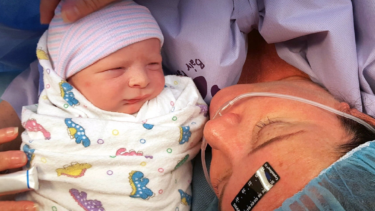A woman lies in a hospital bed wearing a medical gown and oxygen tube, closely holding a newborn wrapped in a patterned blanket. The setting appears to be a hospital or medical facility, with visible medical equipment and attire. The newborn is swaddled in a blanket with colorful animal prints. The overall mood is intimate and tender, capturing a post-birth moment.