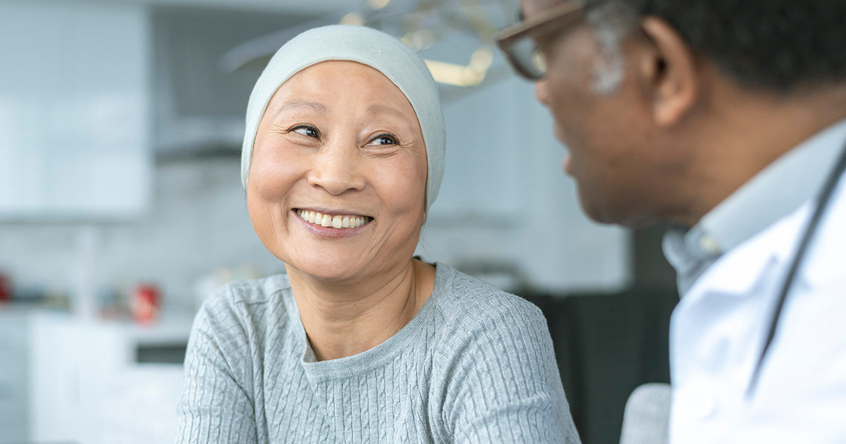 A person wearing a light-colored headscarf and sweater sits across from a healthcare professional in a white coat. The setting appears to be a modern medical office or clinic, with a calm and neutral color palette. The interaction suggests a supportive or informative discussion, emphasizing care and empathy. No visible text or numbers are present in the image.