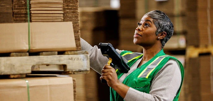 Two warehouse workers in green safety vests discuss tasks in an organized warehouse aisle filed with stacked pallets.
