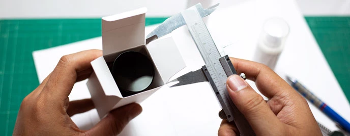 Hands measuring a white cardboard box with a caliper, placed on a table with bottles and design materials in the background.