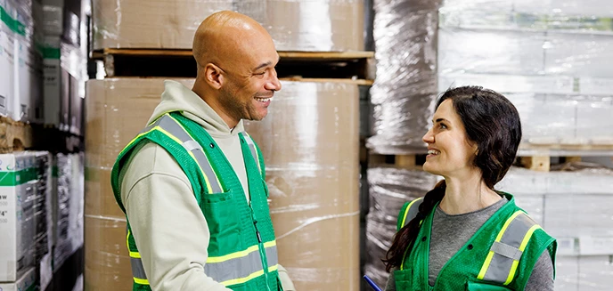 Two warehouse workers in green safety vests discuss tasks in an organized warehouse aisle filed with stacked pallets.