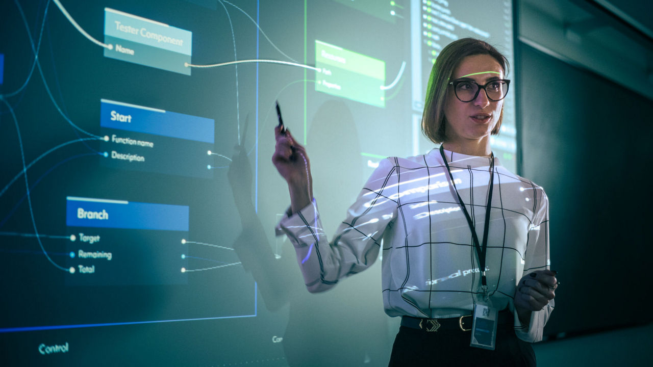 A person stands in a modern classroom or office setting, presenting on a large digital screen.